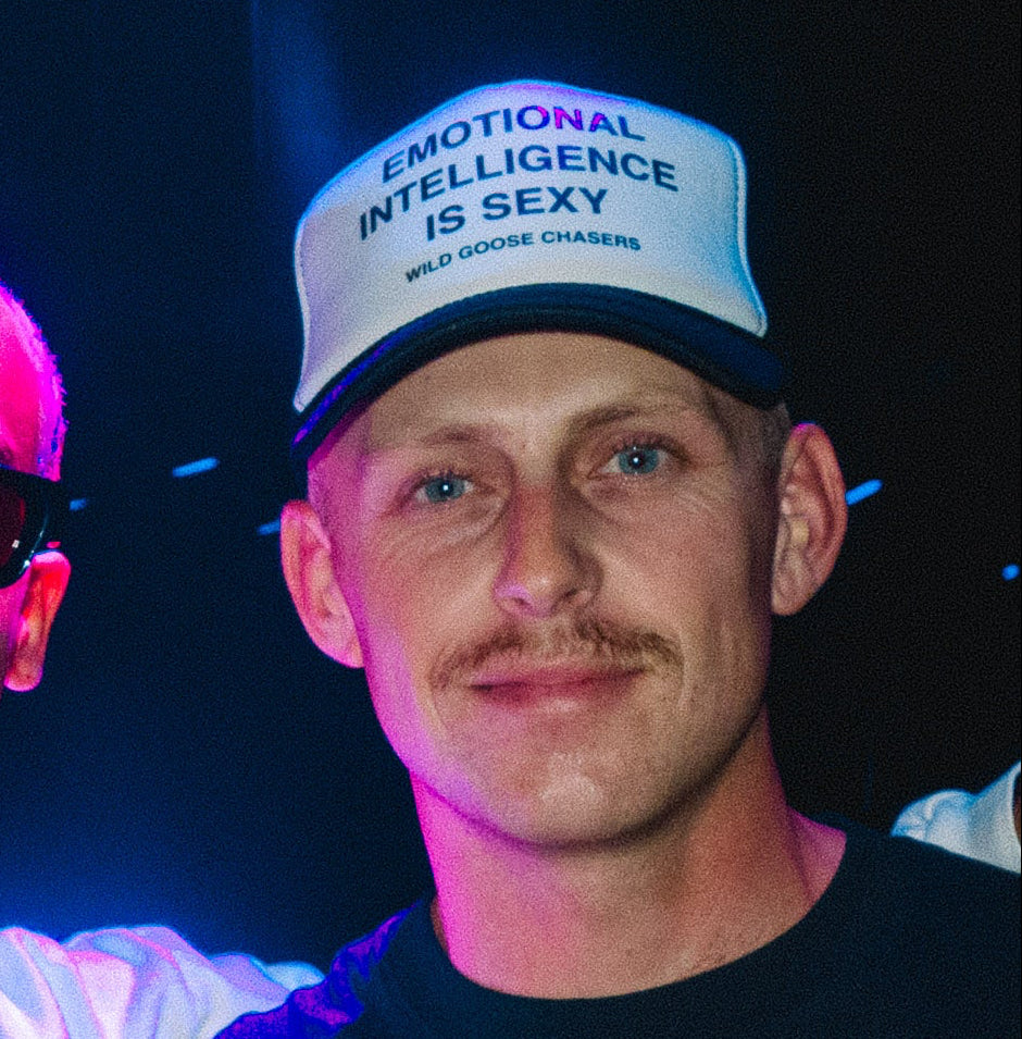 Two men standing close together with one wearing sunglasses and a white shirt, the other wearing a cap and dark clothing.